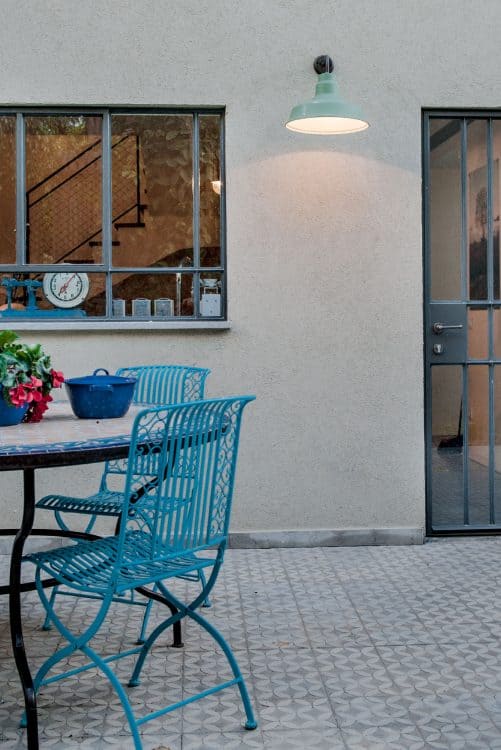 חצר מרוצפת באריחי בטון מוטבעים אפורים חלונות ברזל בלגי כסאות ברזל תכולים ושולחן עם פלטה של פסיפס ממרוקו